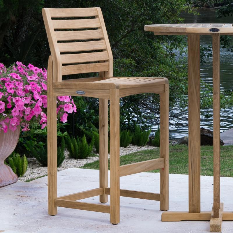 alt=" 70512 Somerset barstool angled next to teak round table with flowering plants and shrubs in background"