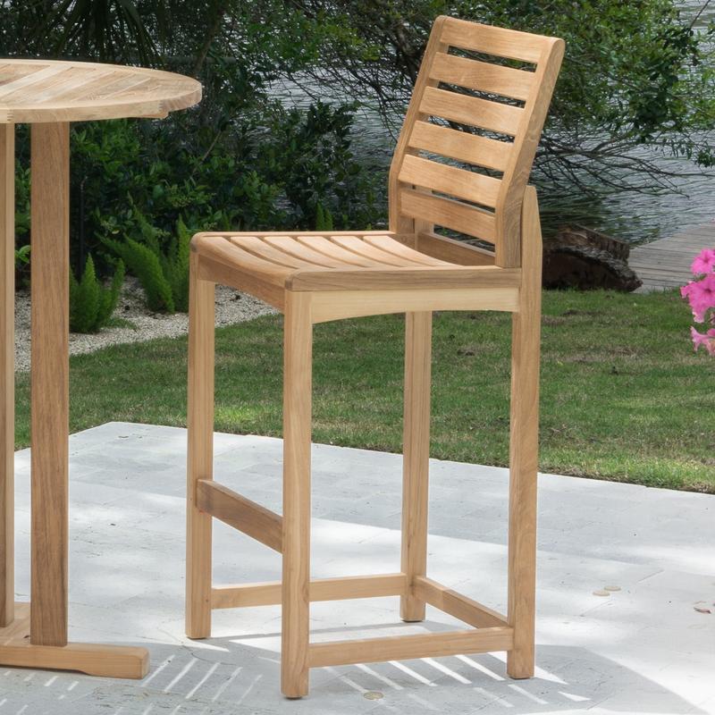 alt="11812 Somerset Side Barstool and 30 inch Teak Bar Table on concrete patio with landscape plants lake and trees in background"