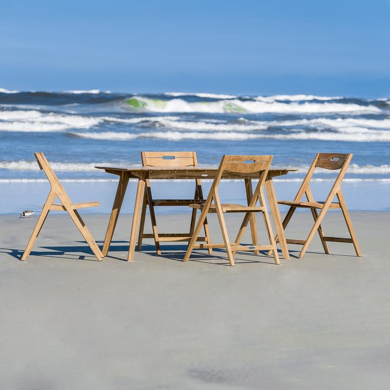 alt="70528 Surf teak 5 piece Dining Set of 4 teak folding side chairs and teak 60 inch rectangular dining table on sand beach overlooking blue ocean and blue sky background "