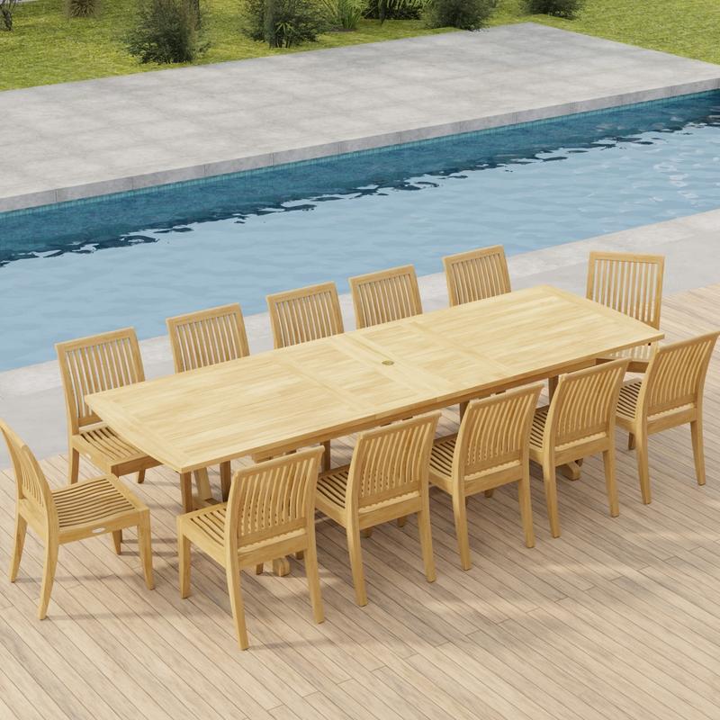 alt="70124 Grand Laguna 13 pc Teak Side Chair Set of Grand Veranda Dining Table and 12 Laguna Side Chairs in angled end view on a patio next to a pool with plants and fence in background"