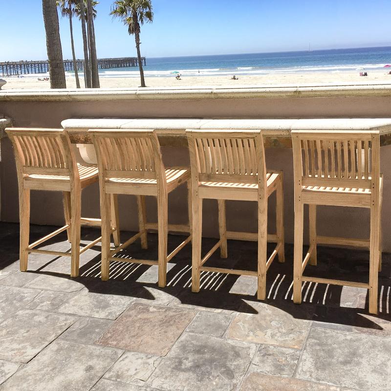 alt="11915 laguna four teak twenty four inch height counter stool on outdoor stone patio against concrete counter looking out to the ocean with palm trees and blue sky background"