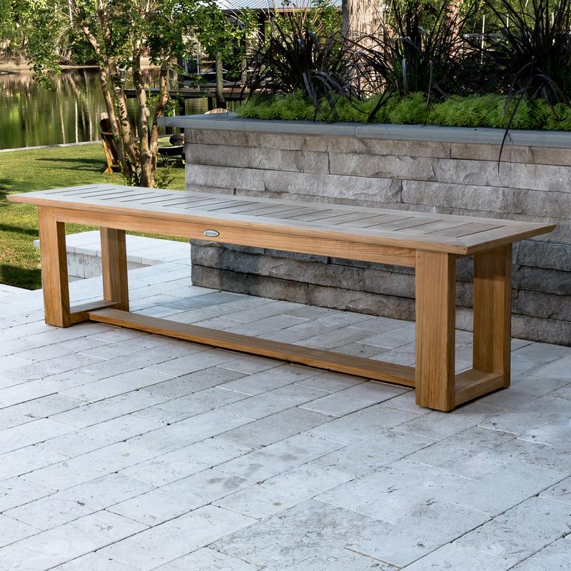alt="13909  Horizon teak 6 foot long Backless Bench with back against raised paver landscape area on concrete stone patio with lake and trees in background"