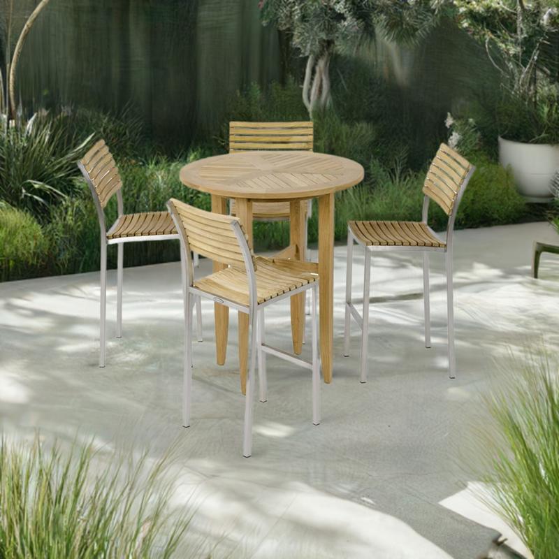 alt="70683 Laguna Round Vogue 5 pc Teak Barstool Set of Laguna 36 inch round teak bar table and 4 Vogue Side Bar Stools angled on concrete patio surrounded by plants with fence in background"