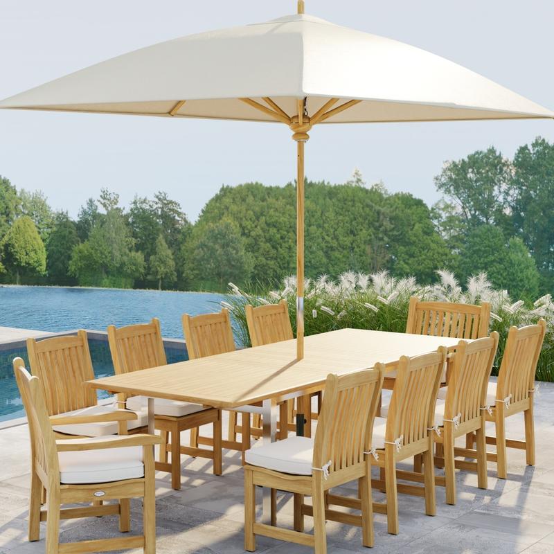 alt="70826  Vogue Veranda 13 pc Teak Dining Set with optional seat cushions and umbrella showing angled end view on patio next to a pool with plants and a lake with trees in background"