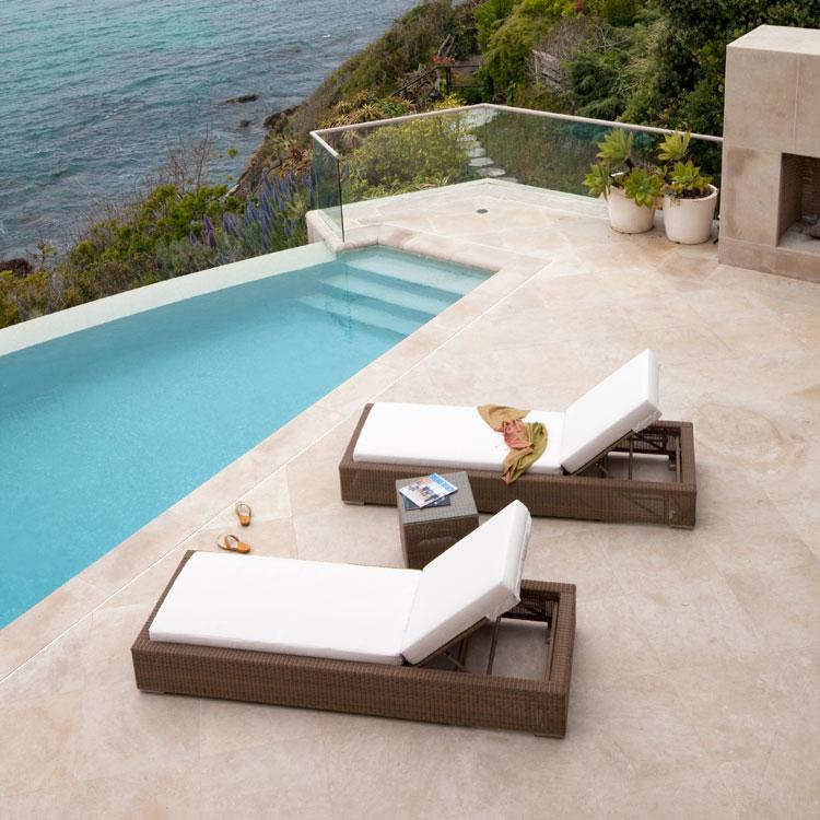 alt="30002dp Malaga two cushioned synthetic wicker chaise loungers sandals between chaises on patio two magazines on side table angled towards pool fireplace glass rail ocean in background"