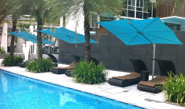 alt="image of SPS2590SFB Spectra umbrella showing 3 in blue canvas on concrete patio by a pool with palm trees and loungers in background"