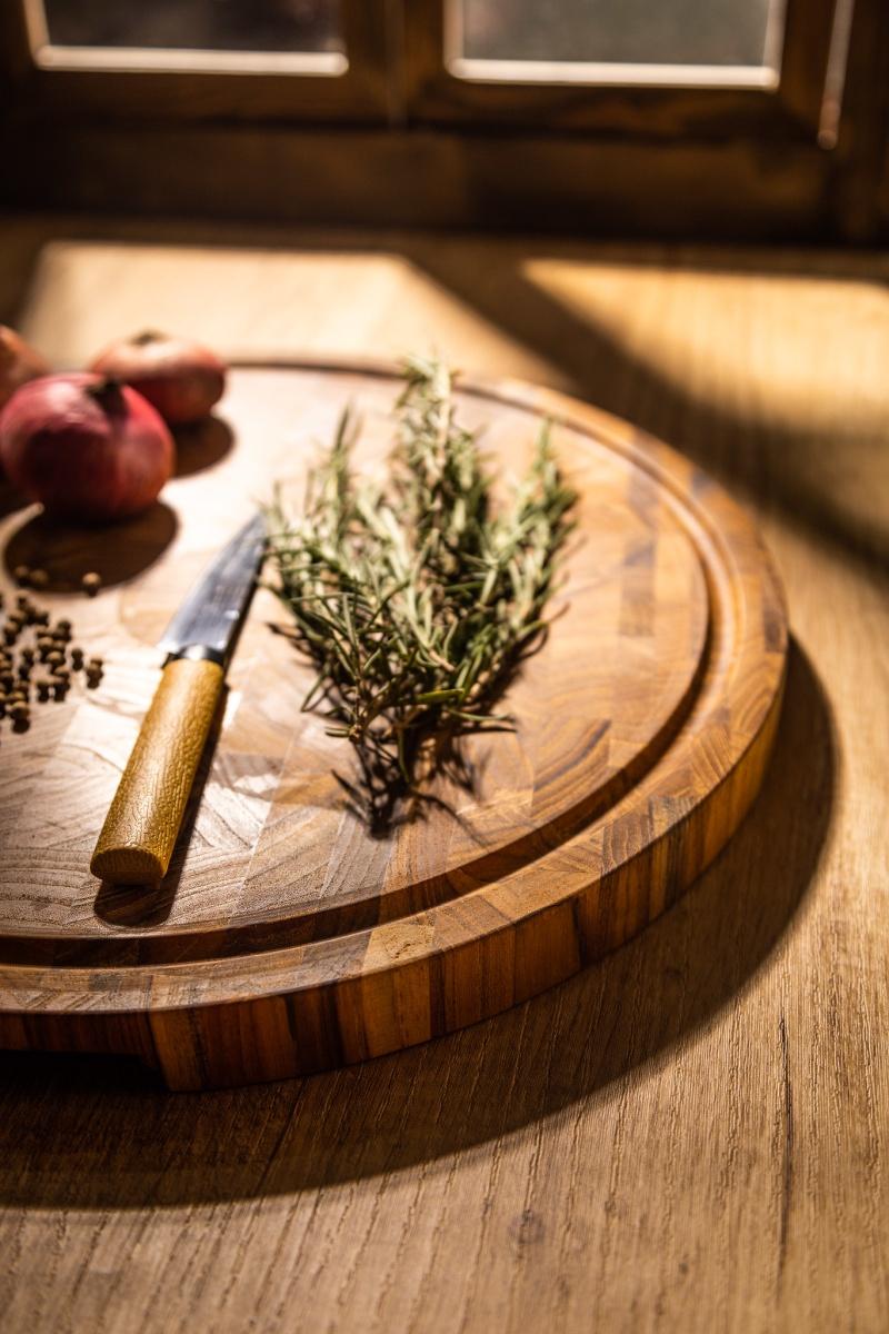 alt="19122 Butcher Block 15 inch Round Charcuterie Cutting Board with rosemary and 3 red onions and whole black pepper and a knife with yellow handle partial aerial view on a counter top and window in background "