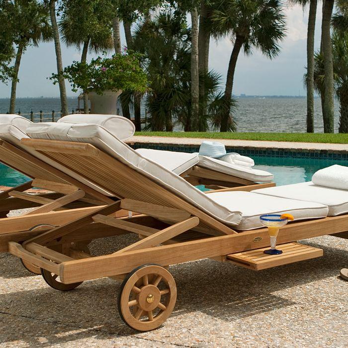 alt="70510 Somerset teak double chaise set with two folded towels and a hat on river rock patio facing pool with palm trees ocean view blue sky in background"