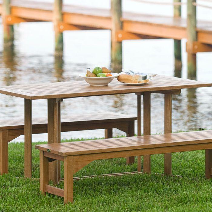 alt="70061 Barbuda Picnic Table teak Dining Set on grass field and white bowl of whole fruit and plate of french bread on table with lake and dock in background"