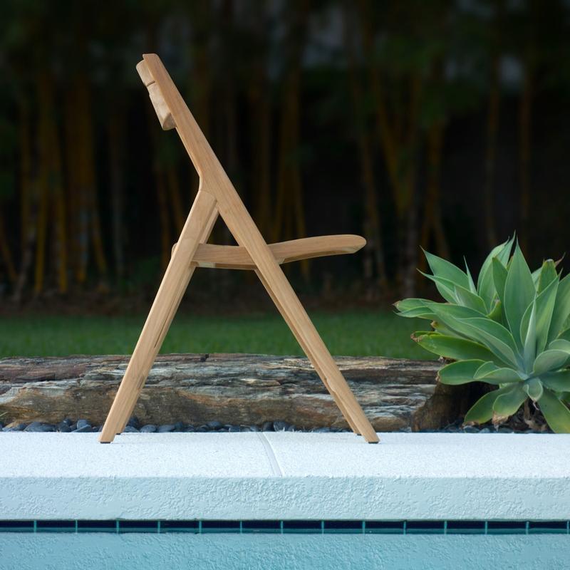 alt="70621 Vogue Laguna teak foldable dining side chair side profile view with pool in front of chair with landscape timber and plant with grass and trees in background"