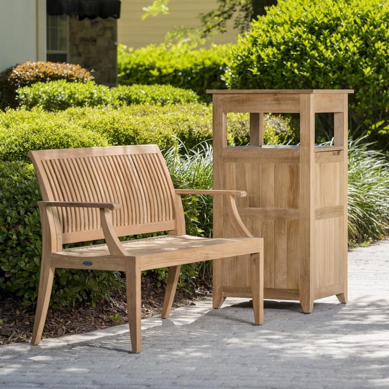 alt="13810 Laguna 4 foot Teak Bench beside teak Palazzo III trash receptacle on concrete walkway with bushes and house in background"