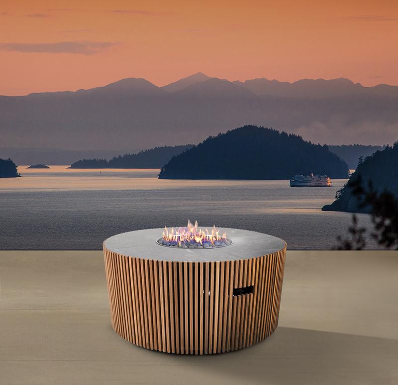 alt="14100 Agora round Fire Table lit in side angled view on outdoor terrace overlooking a boat on a lake with mountains and islands and sunset view in background"