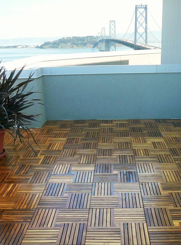 alt="70406 Parquet 10 pack Teak 18 inch Deck Tiles on terrace with a potted plant against a balcony rail with bridge in background"