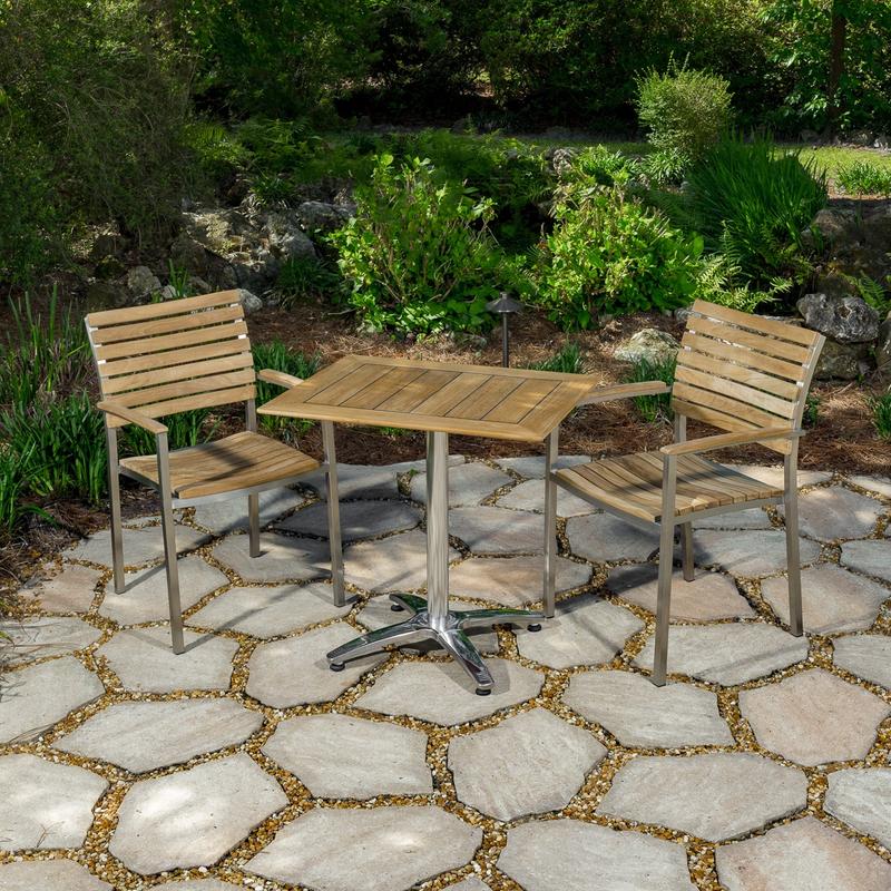 alt="70668 Veranda Vogue Bistro Set of teak and stainless steel rectangular table and 2 teak dining chairs on stone patio with metal sculpture and trees in background "