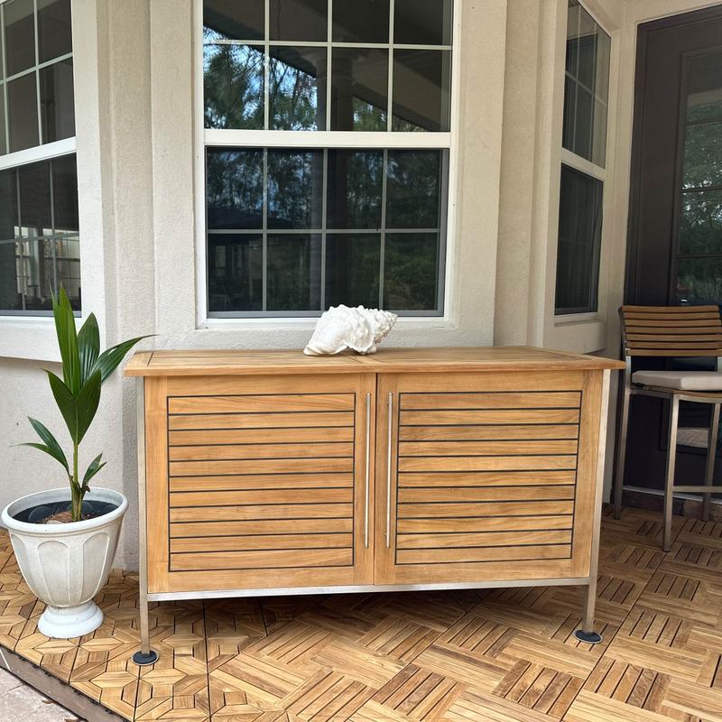 alt="28226 Vogue Teak and Stainless Steel Beverage Console next to a potted plant on teak tiled outdoor patio with windows of a house in the background"