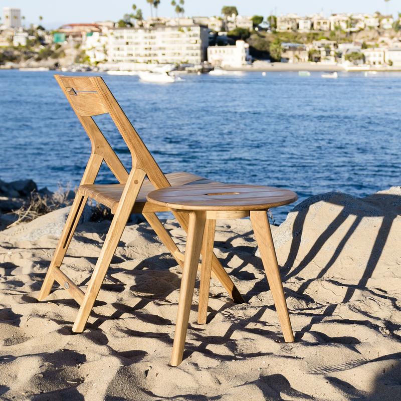 alt="14916 surf teak side table beside surf folding chair on sand by lake with boats and buildings in background"
