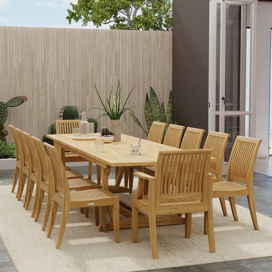 70300 Grand Laguna 13 pc Teak Dining Set with Grand Veranda Dining Table and 12 Laguna Side Chairs on patio next to a house and a fence with plants in the background