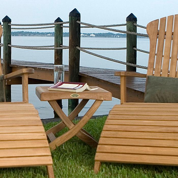 alt="18600 Barbuda Ottoman Side Table with open book face down and glass of water on grass between two adirondack chairs with dock and lake in background"