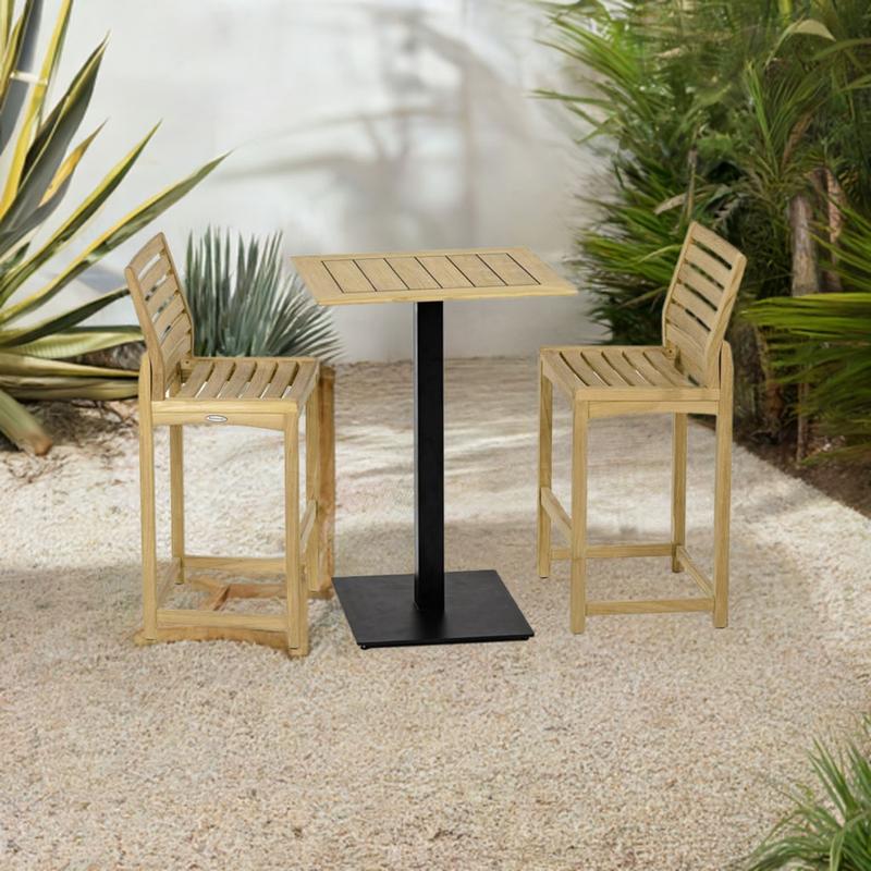 alt="70643 Vogue Somerset 3 pc Barstool Bar Set of Vogue Rectangular teak Table Top on Black Table Base and 2 Somerset Side Barstools angled on stone patio with plants and wall in the background"