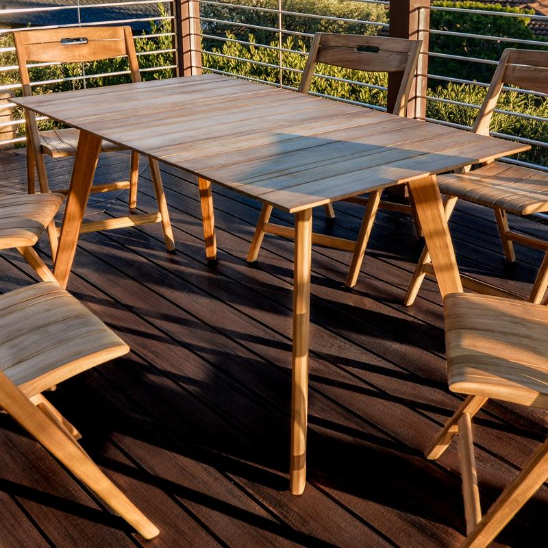 alt="70528 Surf teak 5 piece Dining Set closeup angled view of table on wooden deck with chairs around table with deck railing and shrubs in background"