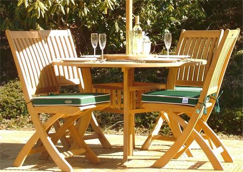 alt="70056 Hyatt Barbuda teak round Dining Set with 4 white plate place settings and 4 wine glasses bottle of wine potted plant on top and optional seat cushions on teak tiles with shrubs in back"