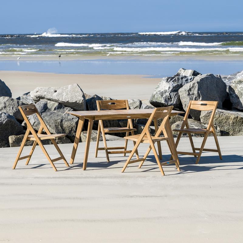 alt="70528 Surf teak 5 piece Dining Set of 4 teak folding side chairs and teak 60 inch rectangular dining table on sand beach with boulders in background overlooking blue ocean"