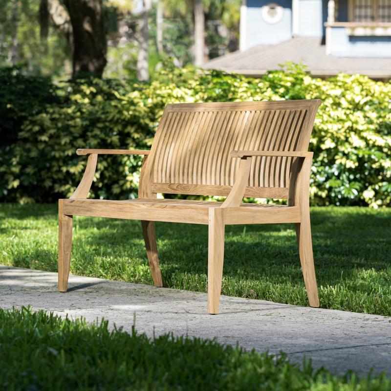 alt="13810 Laguna 4 foot Teak Bench angled side view on concrete walkway with bushes and house in background"