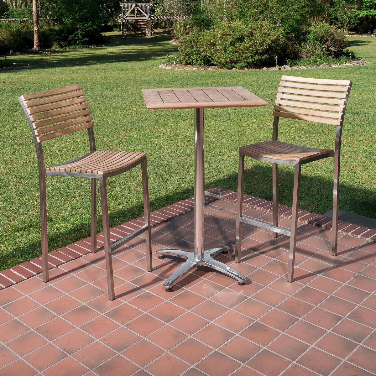 70673 Vogue teak and stainless steel High Bar Set of 2 barstools and 24 inch square teak table on brick paver and tiled outdoor patio with grass and landscape plants in background