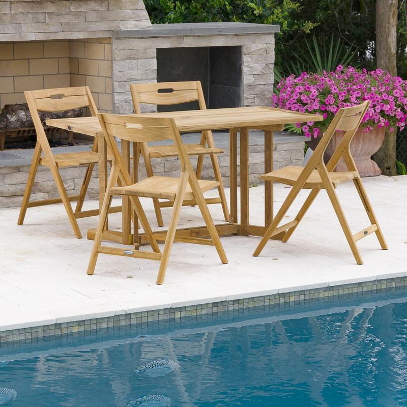 alt="70475 Surf Nevis 5 piece teak rectangular Dining Set on concrete patio and pool with flowering plant next to fireplace trees and shrubs in background"