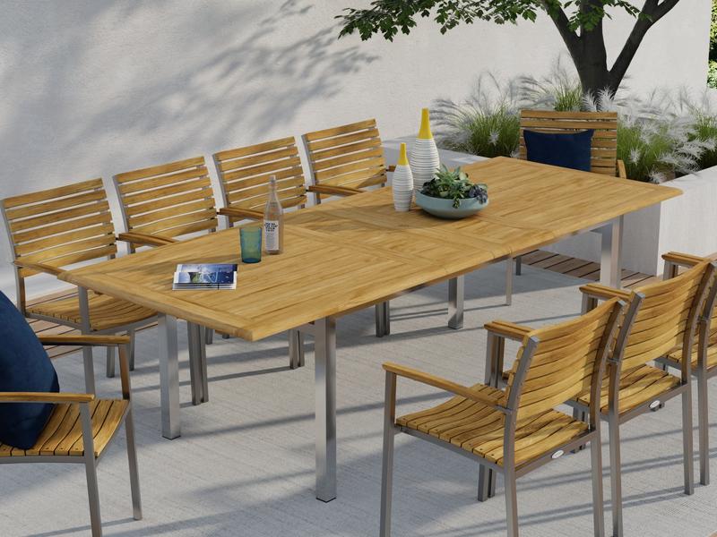 alt="25025 Vogue Extension Table and 9 Vogue Side Chairs angled side view in front of wall on outdoor patio next to a planter box with a tree and landscaping plants in background"