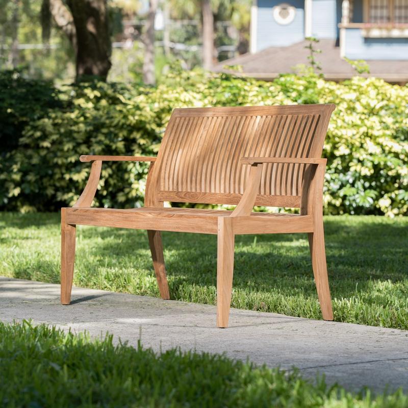 alt="13811ph Laguna teak 6 foot bench on concrete walkway with bushes and house in background"