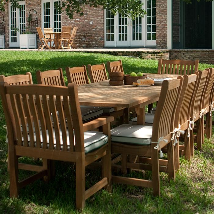 alt="70017 Grand Veranda 13 piece dining set with optional seat cushions bowl of apples plate of bread serving plates and bowls grass lawn brick house round teak dining set in background"
