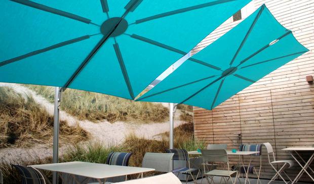 alt="image of SPS2590SFB Spectra umbrella with blue canvas covering over 3 piece bistro sets with hills and building in background"