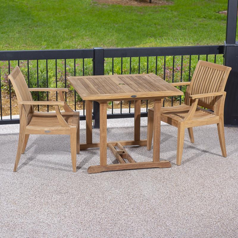 alt="70301 Laguna 3 piece teak Dining Set of a 36 inch square dining table and 2 teak dining armchair on concrete patio against a black railing with grass lawn background"