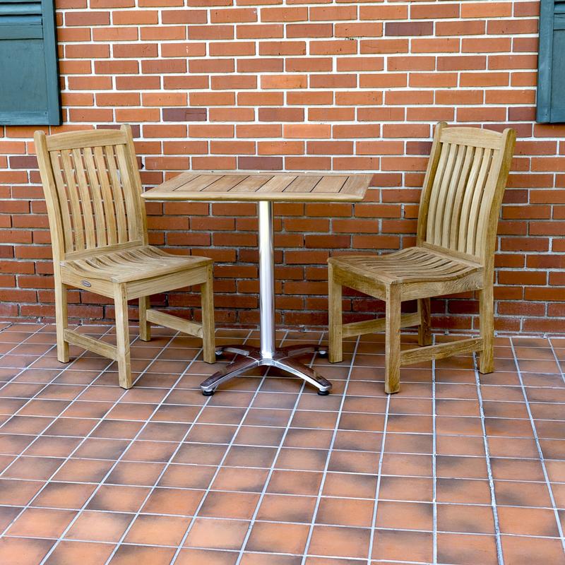 alt="70669 Veranda Vogue Bistro Set of teak and stainless steel rectangular table and 2 teak dining chairs on outdoor tile patio with brick building and green window shutters in background "