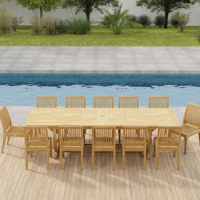 alt="70124 Grand Laguna 13 pc Teak Side Chair Set of Grand Veranda Dining Table and 12 Laguna Side Chairs in side view on a patio in front of a pool with plants and fence in background"