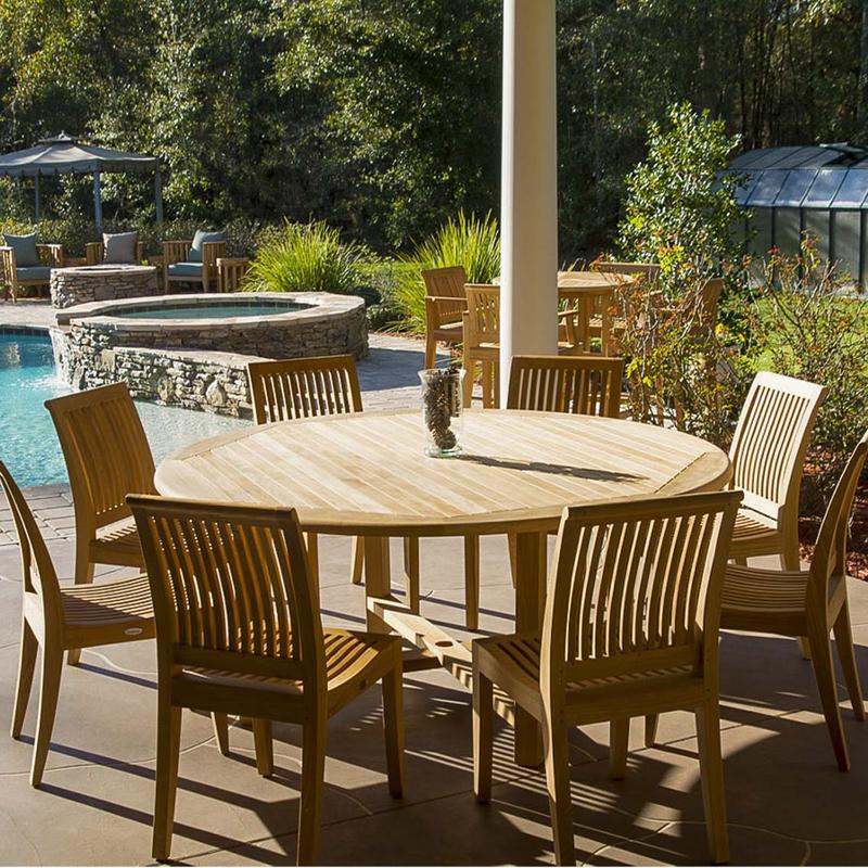 alt="70493 Buckingham Laguna teak 9 piece Dining Set of 8 side chairs and 6 foot round table with vase of pinecones on stone patio with pool in back surrounded by landscape plants "