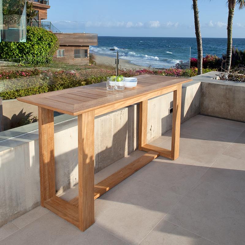 alt="15669 Horizon Teak Console Table angled side view with 2 glasses and bowl of lime and 4 plates on outdoor terrace surrounded by palm trees and landscape plants overlooking ocean"
