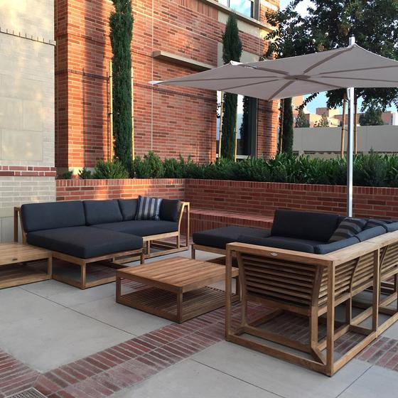 70529 maya eight piece teak lounge set with black cushions market rectangular umbrella set on patio with brick planter with plants trees and brick building background