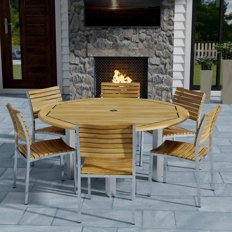alt="70439 Vogue Dining Set for 6 of Vogue Teak and Stainless Steel 60 inch Round Dining Table and 6 Vogue Side Dining Chairs angled on patio with an outdoor fireplace in the background "