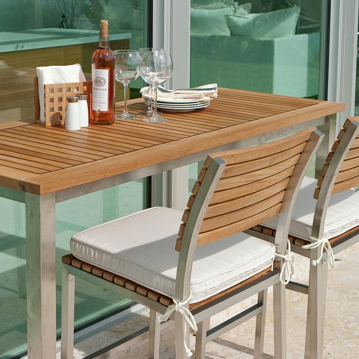 alt="72000MTO Vogue teak and stainless steel bar set with seat cushions and 2 wine glasses and wine bottle and condiments and napkins on stone patio against glass wall closeup angled view"