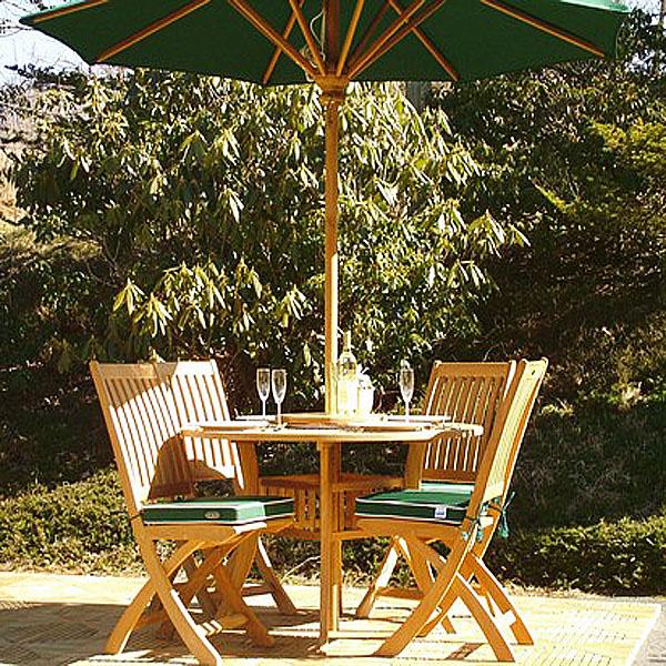 alt="70056 Hyatt Barbuda teak 5 piece Dining Set showing 4 place settings and wine glasses bottle of wine on table with optional open market umbrella on teak tiles with shrubs in background"