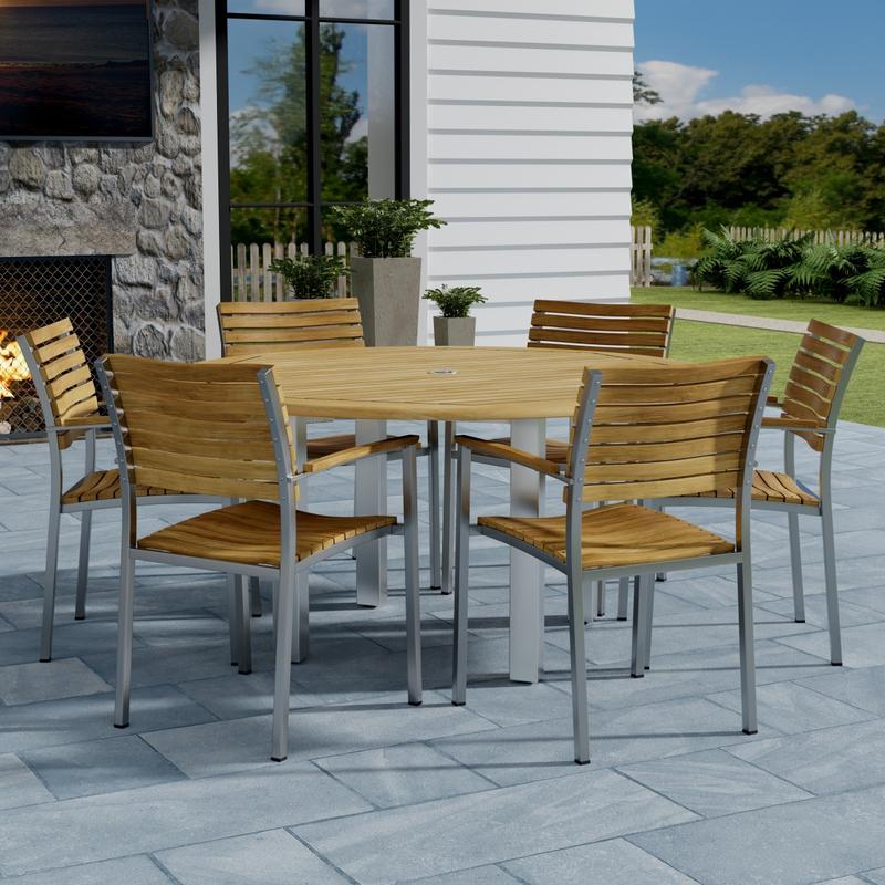 alt="70442 Vogue Teak 7 pc Dining Set showing Vogue 60 inch Round Dining Table and 6 Dining Chairs side view on patio with window and fence with trees in background"