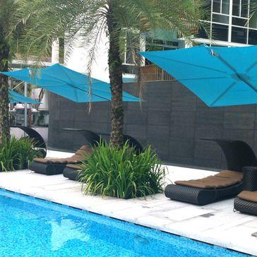 alt="image of SPS2590SFB Spectra umbrella showing 3 in blue canvas on concrete patio by a pool with palm trees and loungers in background"