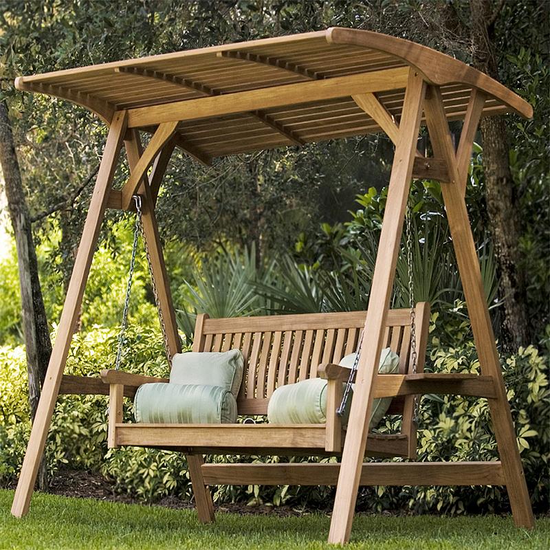 alt="13955 veranda swinging bench and teak canopy with four throw pillows on grass angled with trees and shrubs in background"