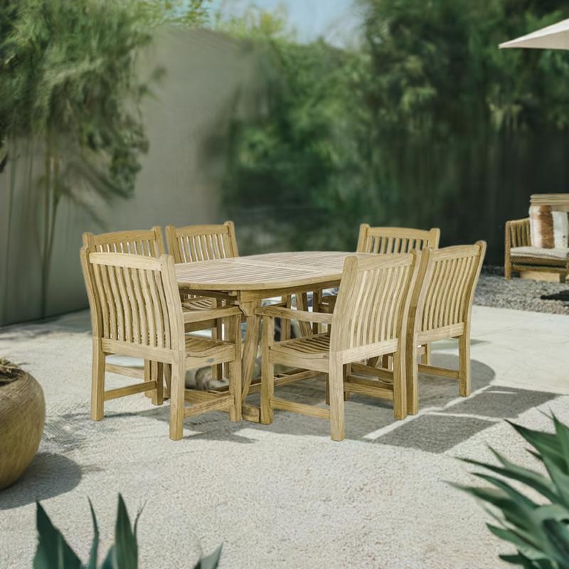 alt="70002 Montserrat 7 piece teak oval Dining Set of Monsterrat Oval Teak Dining Table and 6 Veranda Dining Chairs angled on rock patio surrounded by plants with privacy wall in background"