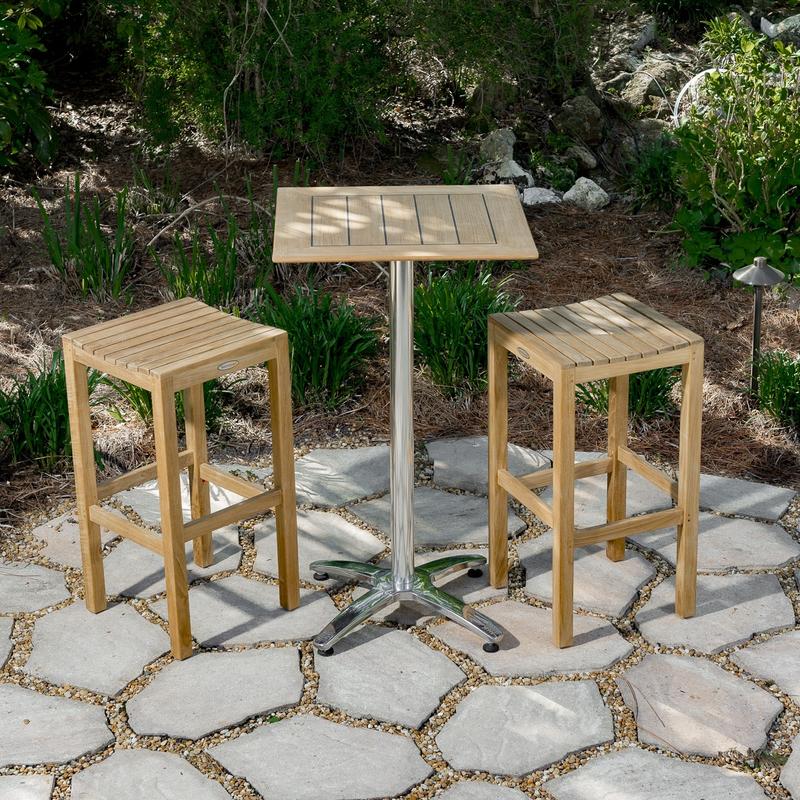 alt="70670 Somerset teak Pub Bar Table and 2 teak Backless Barstools on stone patio with landscape shrubs and trees in background"