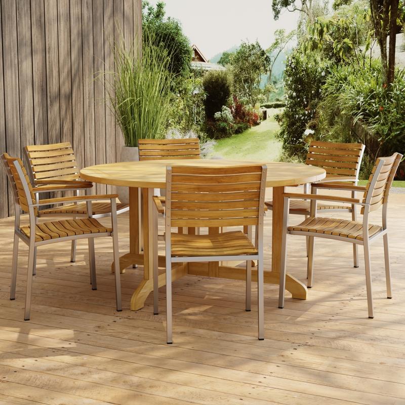 alt="70349 Barbuda Vogue Teak 7 pc Dining Set in side view on paver patio with wood privacy wall with landscaping plants and trees in background"