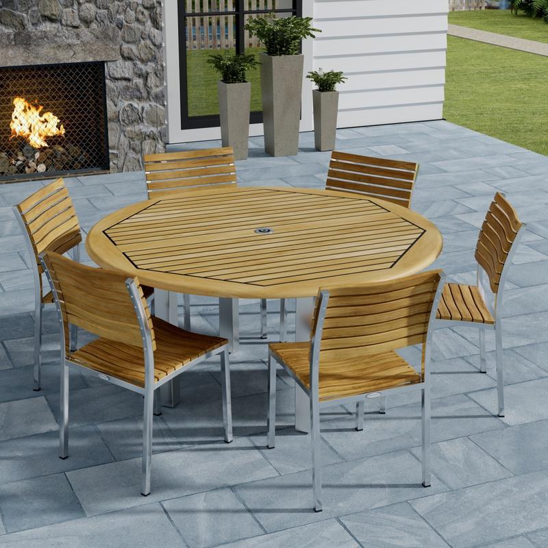 alt="70439 Vogue Dining Set for 6 of Vogue Teak and Stainless Steel 60 inch Round Dining Table and 6 Vogue Side Dining Chairs angled aerial view with an outdoor fireplace in the background "