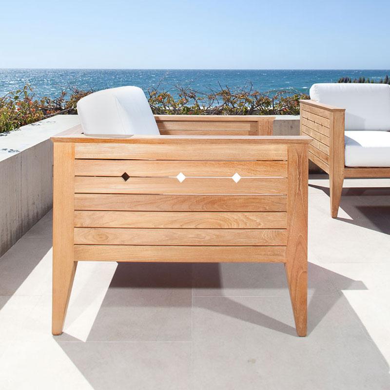 alt="12160dp Craftsman cushioned teak armchair outside on concrete terrace with flora and ocean background"